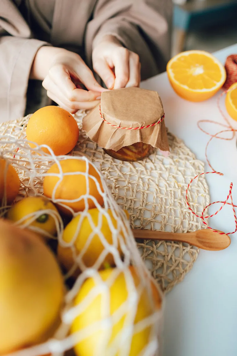 A jar of marmalade being tied with paper and string, oranges in a string bag beside it